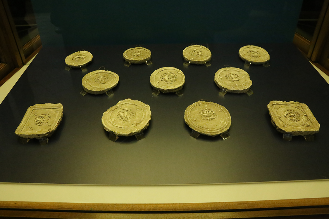 LoScrignoDelCardinale_63.jpg - La bacheca contiene 11 calchi in gesso che riproducono alcuni medaglioni medievali di Limoges --  già decoranti un cofano di Guala Bicchieri — approdati nel XVI secolo a Biella e reimpiegati per impreziosire il coro rinascimentale della chiesa. Vennero realizzati a scopo didattico intorno alla fine dell’Ottocento nei laboratori delle Scuole Professionali di Biella, dal 1873 ospitate nei locali del convento di San Sebastiano. Si tratta di calchi positivi, prodotti senza staccare gli originali dai dossali del coro. Il calco con figura di arpia (prima fila a sinistra, terzo in basso) è il calco del medaglione OA 9475 del Louvre (esposto nella sezione 2, vetrina sud). Nella seconda metà del XIX secolo, nel generale clima di riscoperta del Medioevo, le Scuole di arti e mestieri incentivavano lo studio delle tecniche artistiche medievali, come lo smalto champlevé.